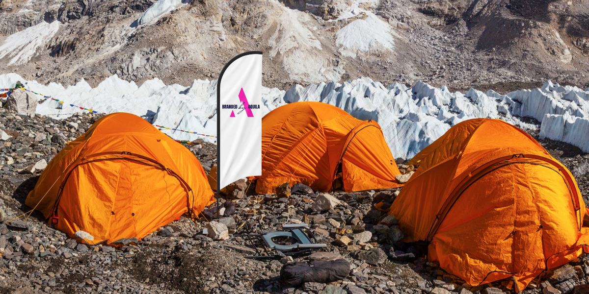 Three orange tents at the base of a mountain with a BbA flag next to them